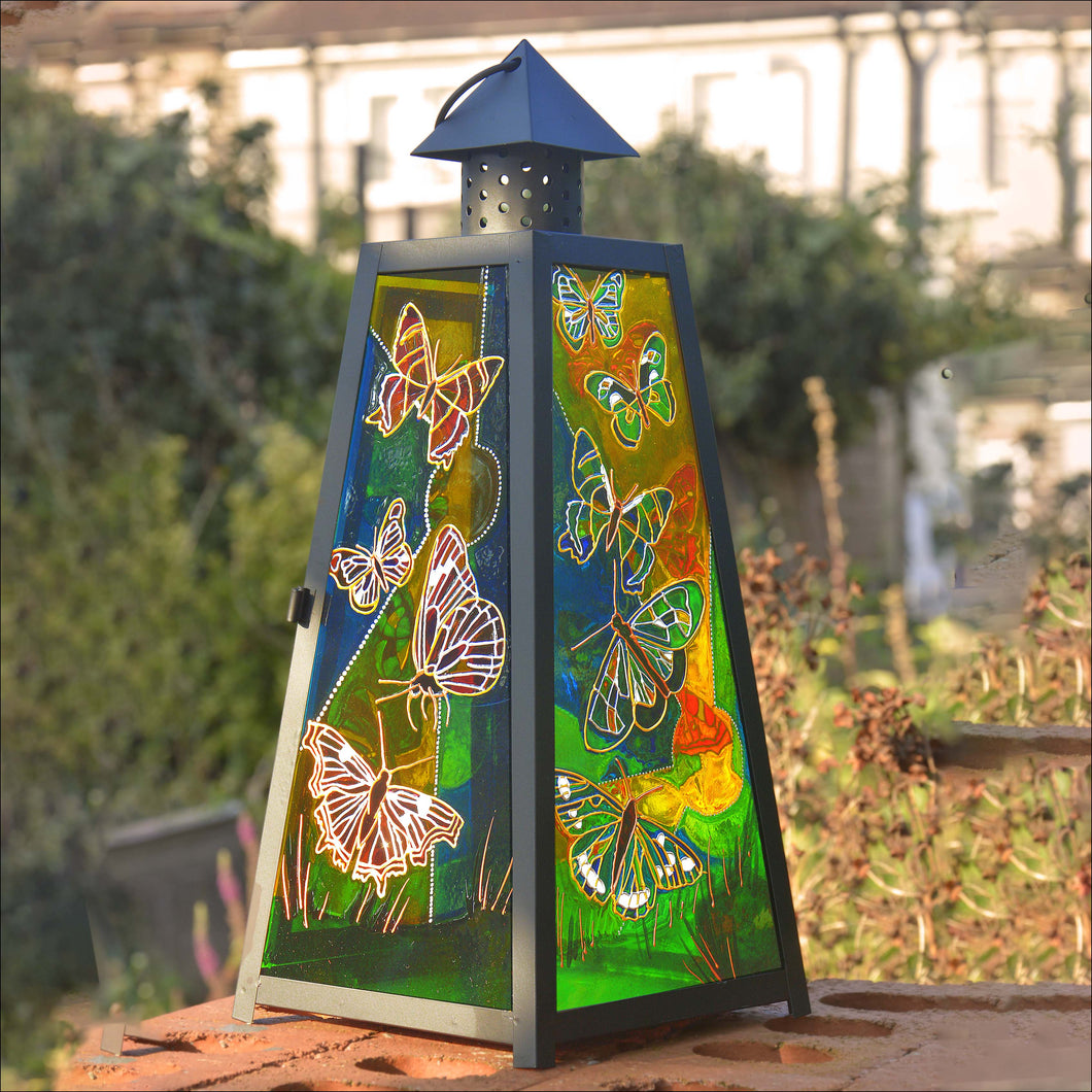 A large black pyramid shaped candle lantern by Ornately Lanterns hand painted with butterflies in a garden lit by sunshine