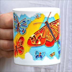 A hand holds a white ceramic coffee mug, printed with a summer design of blue sky, sunshine & lots of colourful butterflies