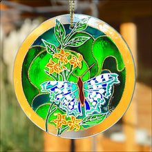 A stained glass circle suncatcher with a large blue & silver butterfly against golden jasmine on a green patterned background