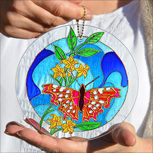Two hands hold a stained glass suncatcher painted in shades of blue with a pink and red butterfly alighting on yellow jasmine