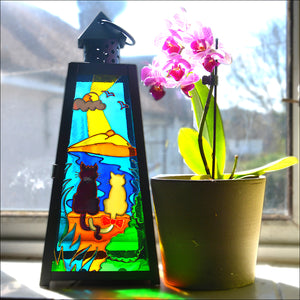 Glowing stained glass candle lantern painted to show cats on a beach by the ocean – it sits on a windowsill next to orchids