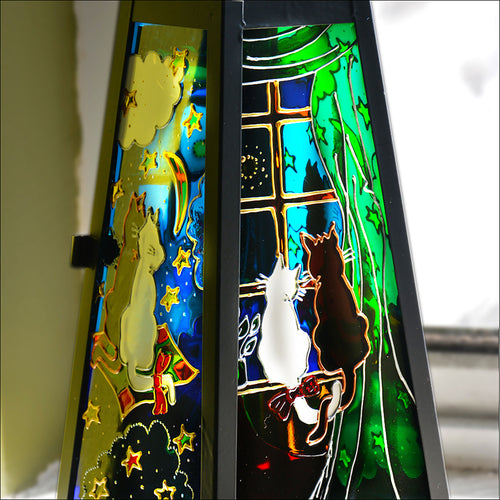 A stained glass candle lantern shows a cat couple sitting at the window – and then travelling through the night sky