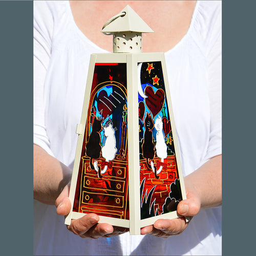 Two hands hold a shabby chic cream candle lantern with stained glass paintings of a romantic cat couple in glowing colours