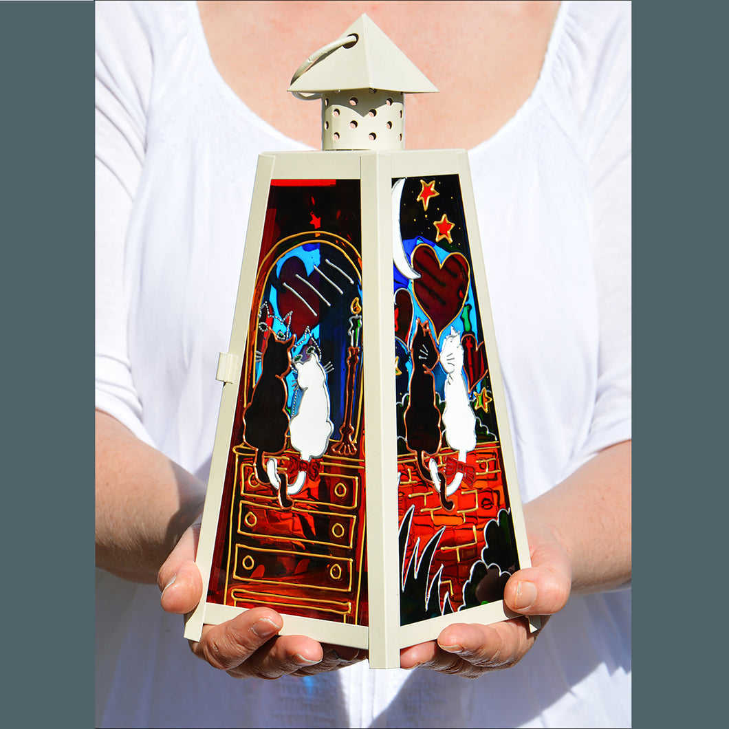 Two hands hold a shabby chic cream candle lantern with stained glass paintings of a romantic cat couple in glowing colours