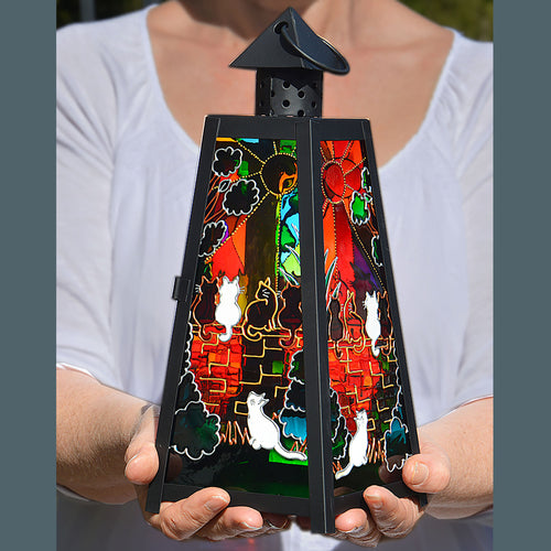 Two hands hold a candle lantern featuring stained glass panels - five cats sitting outdoors on a wall in sunshine & at sunset