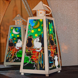 A small cream tealight lantern features stained glass panels of cats on a garden wall in sunshine & under a starry night sky
