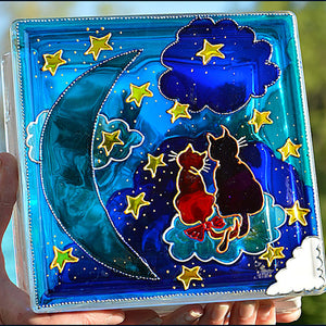 Two hands hold a glass block garden suncatcher painted in blue with two cats amongst the stars on a moonlit night