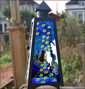 A stained glass candle lantern in a garden features painted panels of a cat couple sitting on a blue moon amongst the stars