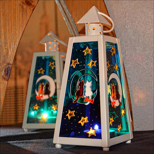 A small cream tealight lantern features stained glass panels of a cat couple sitting on the moon amongst stars in a night sky