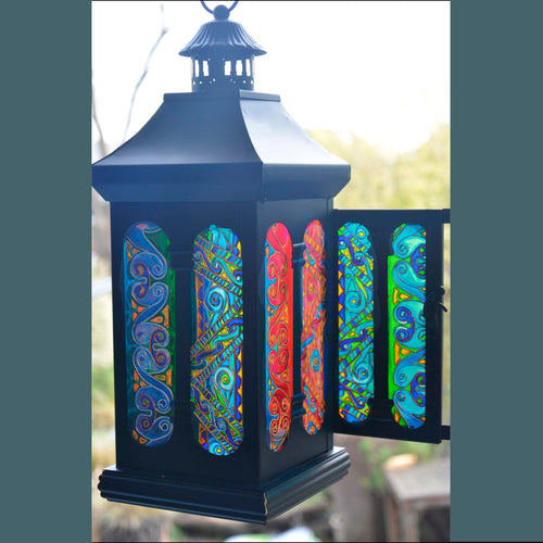 A black metal candle lantern with stained glass panels patterned with intricate Celtic Knot designs in blue green, red & pink