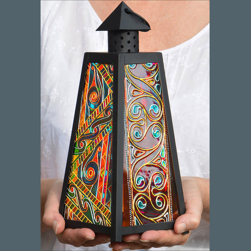 Two hands hold a medium size candle lantern with colourful stained glass panels painted with an intricate Celtic Knots design