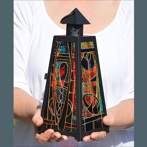 Two hands hold a stained glass candle lamp delicately hand painted with a colourful Charles Rennie Mackintosh style design