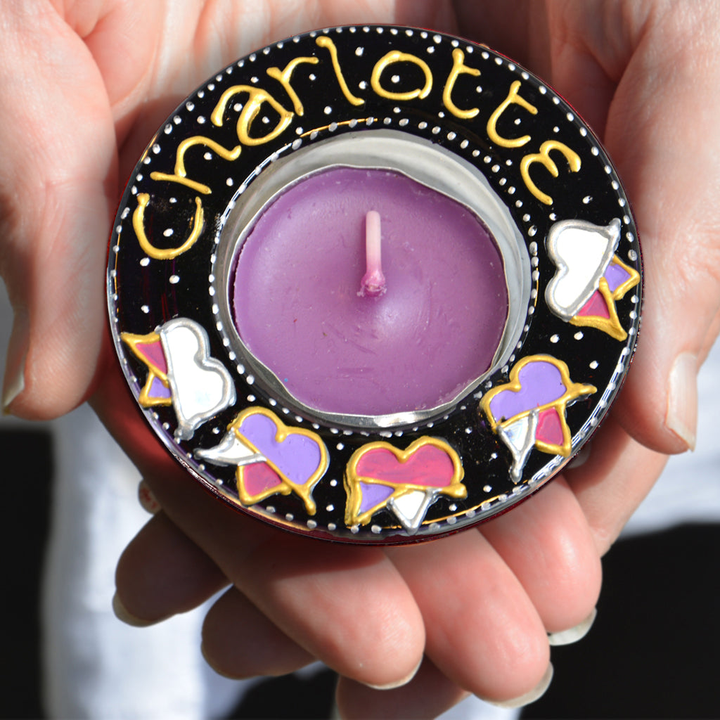 Two hands hold a personalised tealight holder hand painted in dark purple with radiant tulips and the name Charlotte in gold