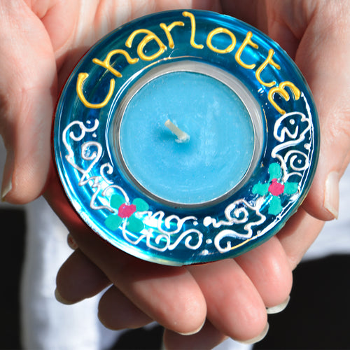 Two hands hold a personalised tealight holder hand painted in sky blue with tiny green daisies and the name Charlotte in gold
