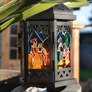 A hand-painted candle lantern shows six Christmas camels walking through a glowing golden desert against a vivid blue night sky
