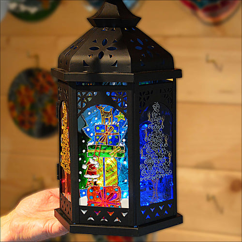 A black metal holiday lantern hand painted with colourful Christmas trees and a snowy Santa climbing a pile of festive gifts