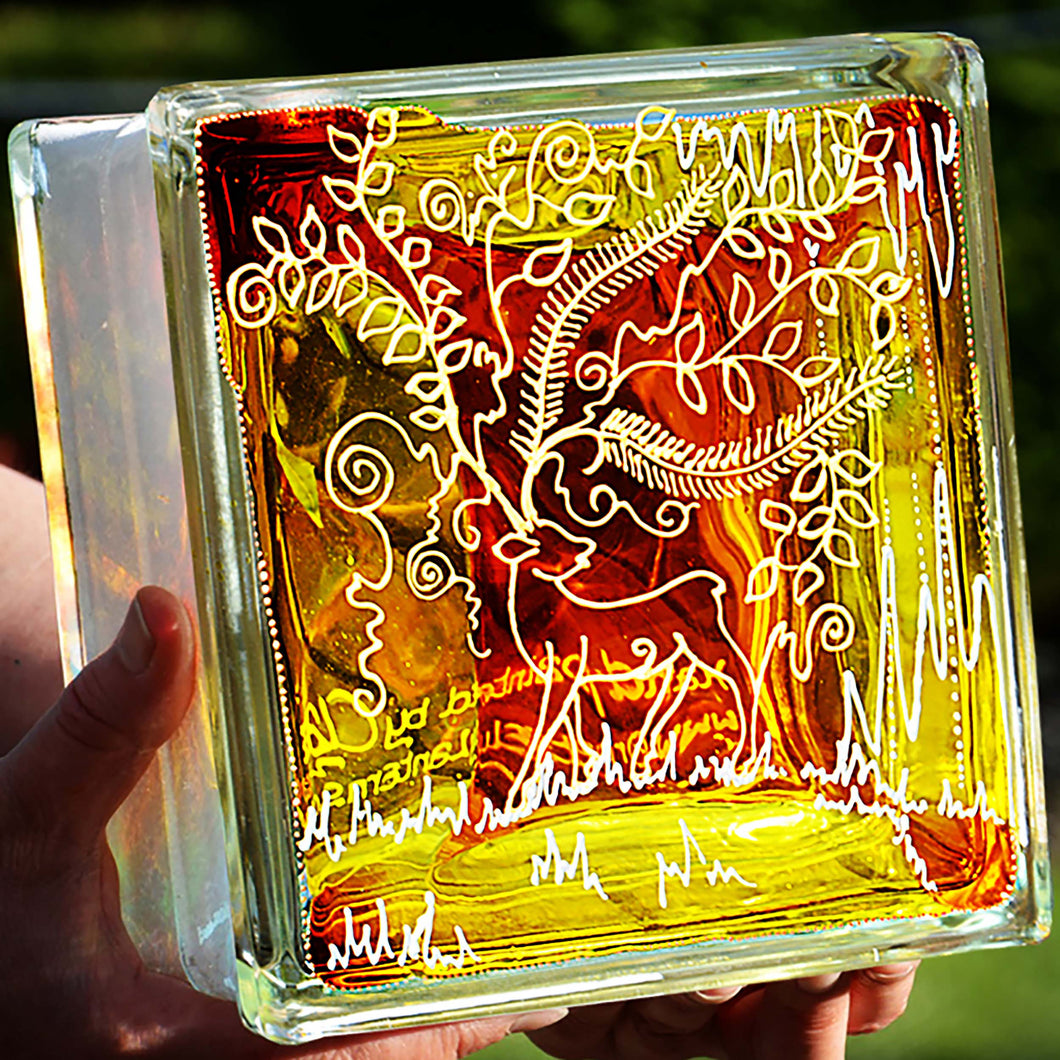 Two hands hold a stained glass block painted in bright yellow and gold with a Christmas reindeer and his lacy floral antlers
