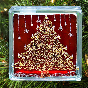 A stained glass cube hand painted with a lacy swirling gold Christmas tree, silver snowflakes and stars against glowing red