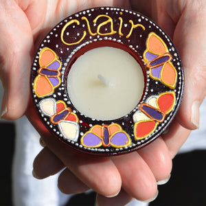 Hands hold a red stained glass tealight holder, hand painted with colourful butterflies and personalised with the name Clair
