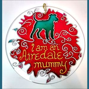 A six inch hanging glass suncatcher, hand painted in pink & silver, with a turquoise dog & the text “I am an Airedale mummy”
