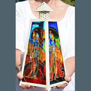 Two hands holding a large pyramid shaped cream candle lantern with stained glass panels with two colourful Art Deco ladies