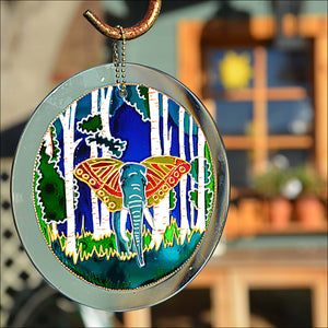 A glowing stained glass suncatcher is painted with a forest and a fantasy blue elephant with golden butterfly wings as ears