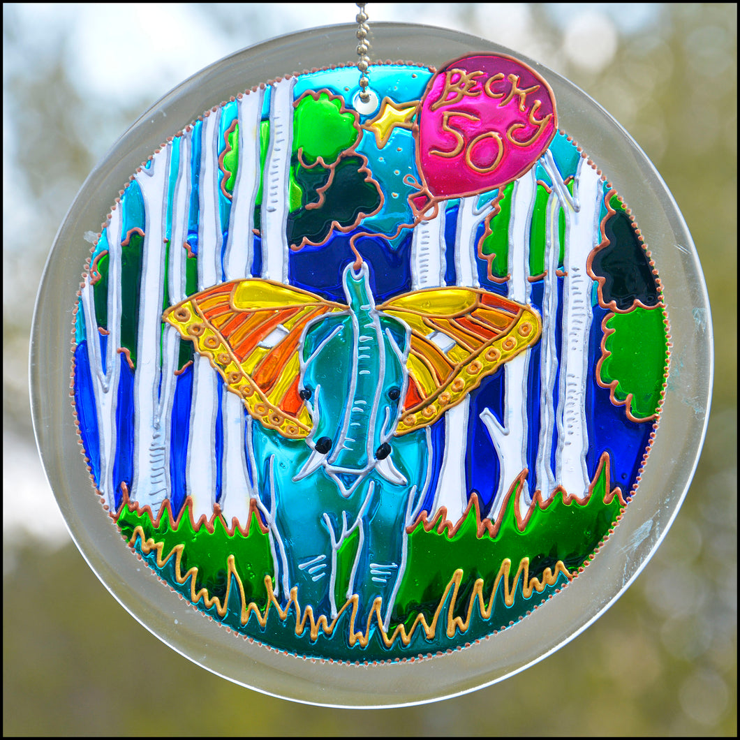 A hanging stain suncatcher personalised to celebrate a birthday, painted with a blue elephant with golden butterfly wing ears