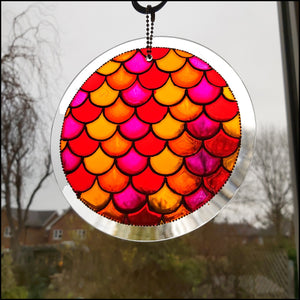 A stained glass painting on a circular hanging suncatcher: abstract fish scales in sunset colours – gold, pink, red, orange