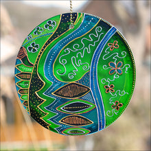 A stained glass suncatcher original painted with an ornate abstract folk art pattern in glowing shades of green and turquoise