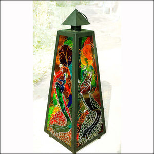 A tall stained glass candle lantern with an elegant art deco style lady riding a colourful peacock shown on all 4 panels