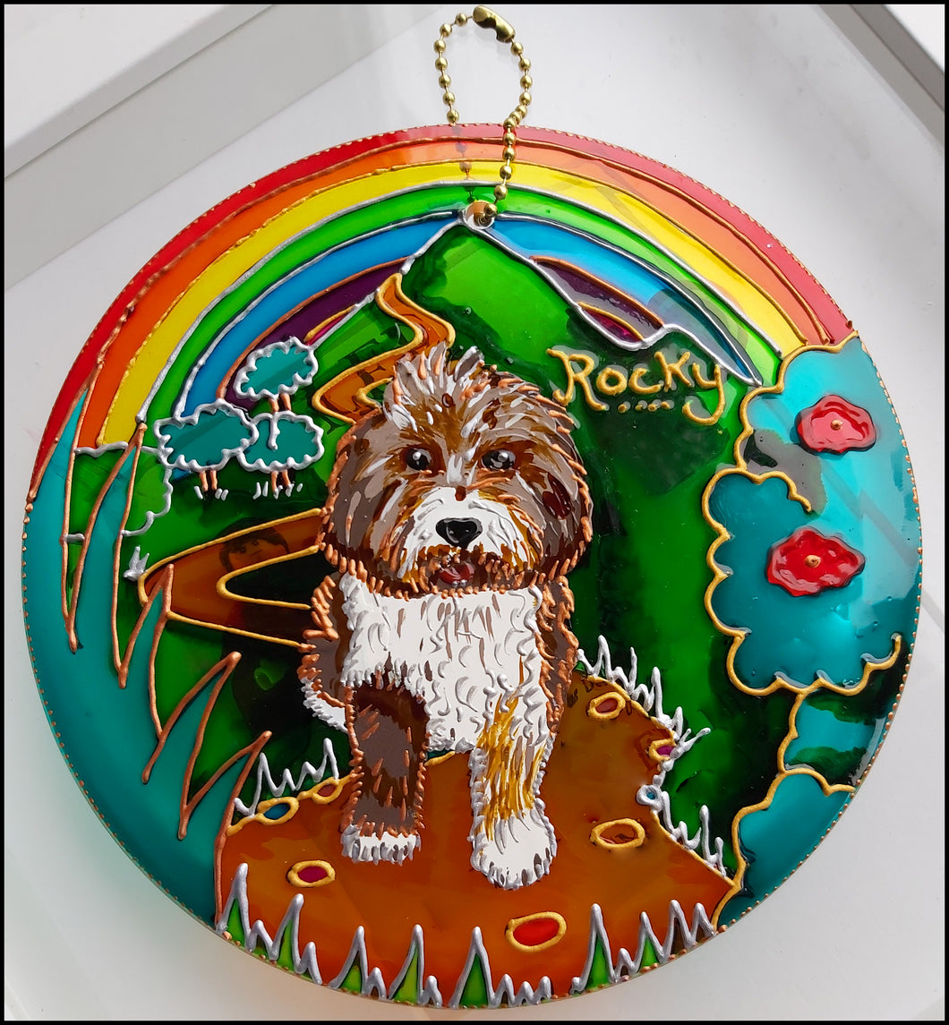 A hanging glass suncatcher pet memorial hand painted with Rocky, a Tibetan Terrier, on a mountain path with a rainbow behind