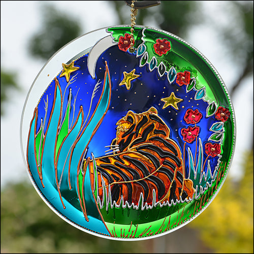 A stained glass suncatcher hand painted in glowing colour with a golden tiger lying in the green grass under moon and stars