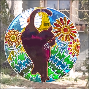 A black witches cat poses against the green grass and big colourful flowers of a sunny garden on a glass circle suncatcher